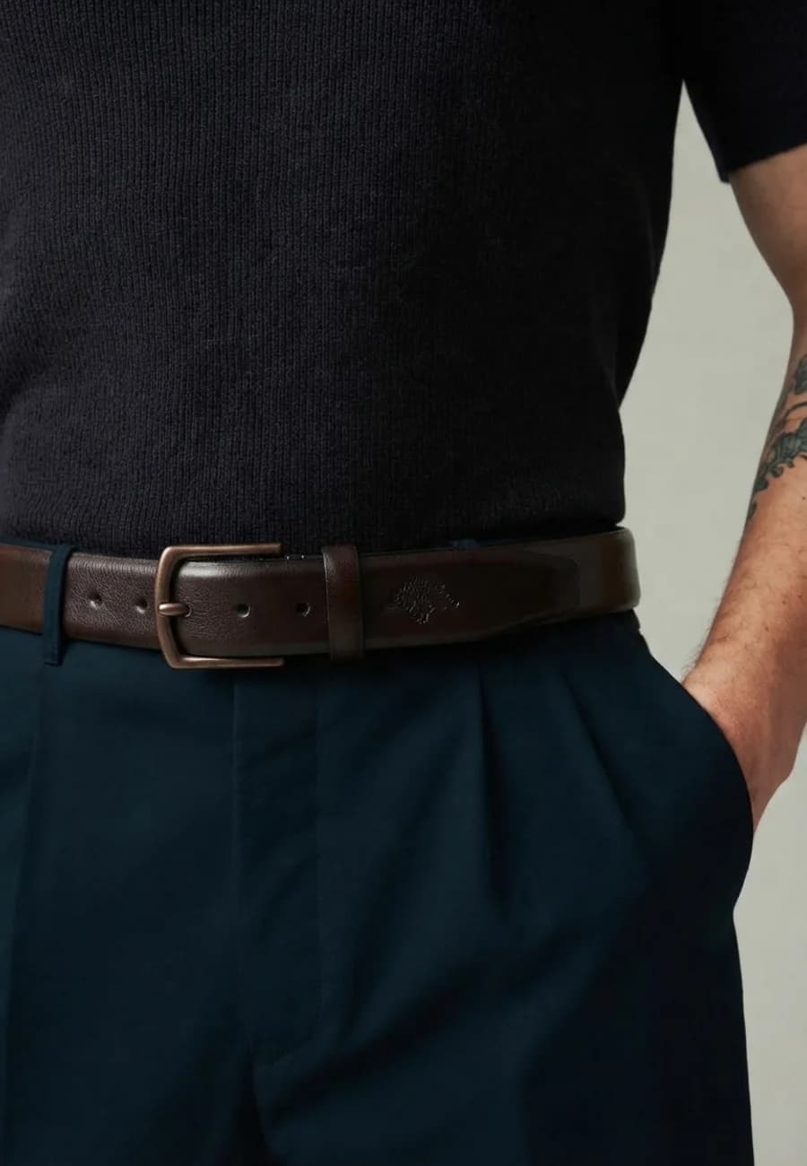 Men's dark brown leather belt with a bronze buckle worn with navy pleated trousers and a navy polo
