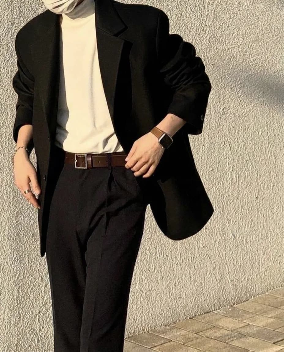 Black blazer worn open over a cream turtleneck, with black trousers, a brown belt, and a brown-strap watch