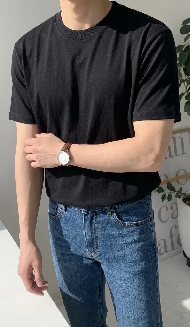 Man in a black crew-neck T-shirt and mid-blue jeans, wearing a round watch with a brown leather strap