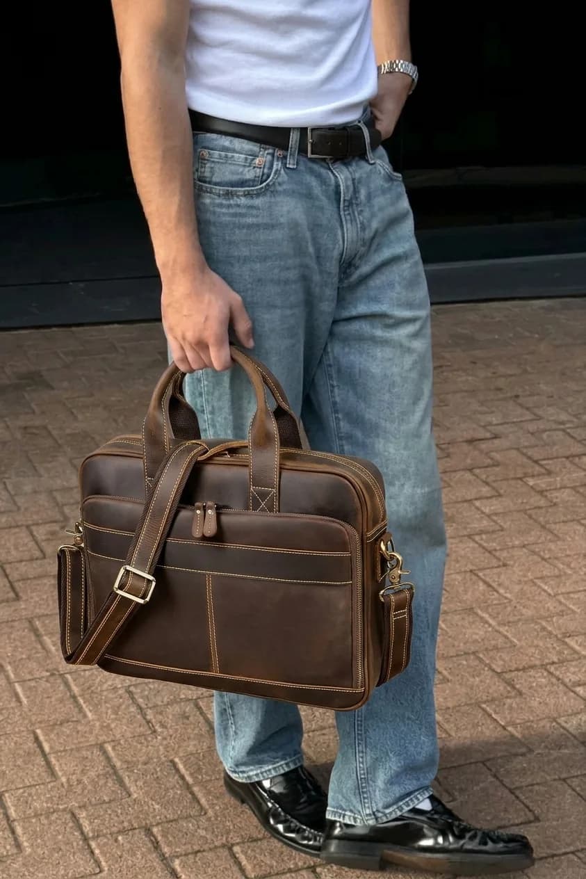 Brown leather briefcase with top handles and a shoulder strap, carried with jeans and black loafers