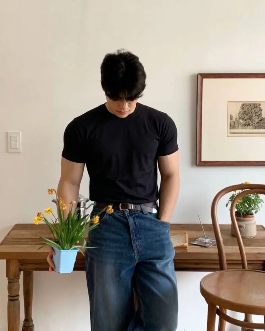 Man in a black crew-neck T-shirt, blue jeans, and a brown belt, holding a small pot of yellow flowers
