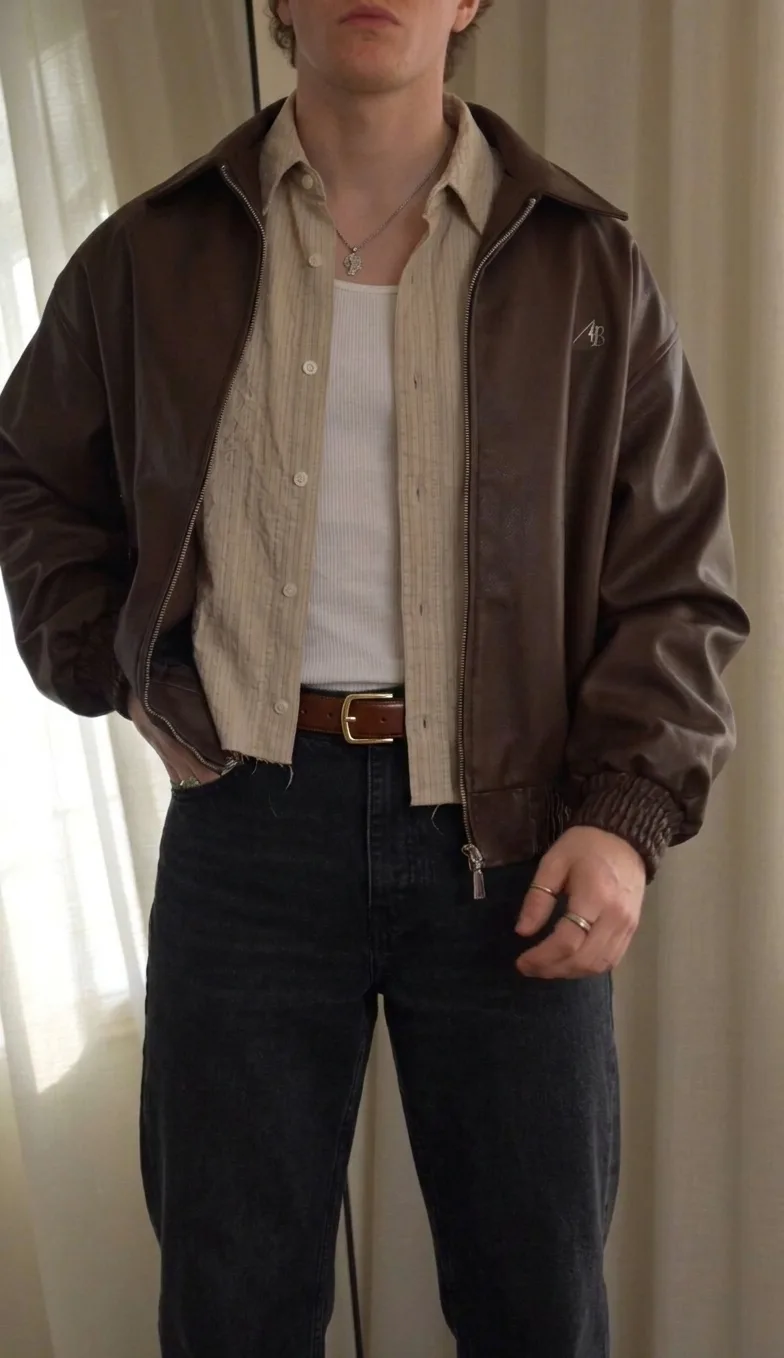 Man in a brown leather jacket over a striped shirt and white tank, with black jeans and a brown belt