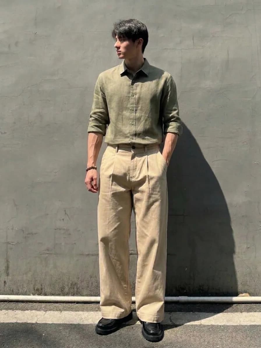 Man in an olive button-up shirt and beige pleated trousers with black shoes, standing against a concrete wall