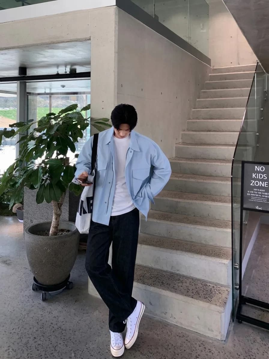 Man in a light blue overshirt over a white T-shirt, black jeans, and white sneakers, standing by a concrete staircase