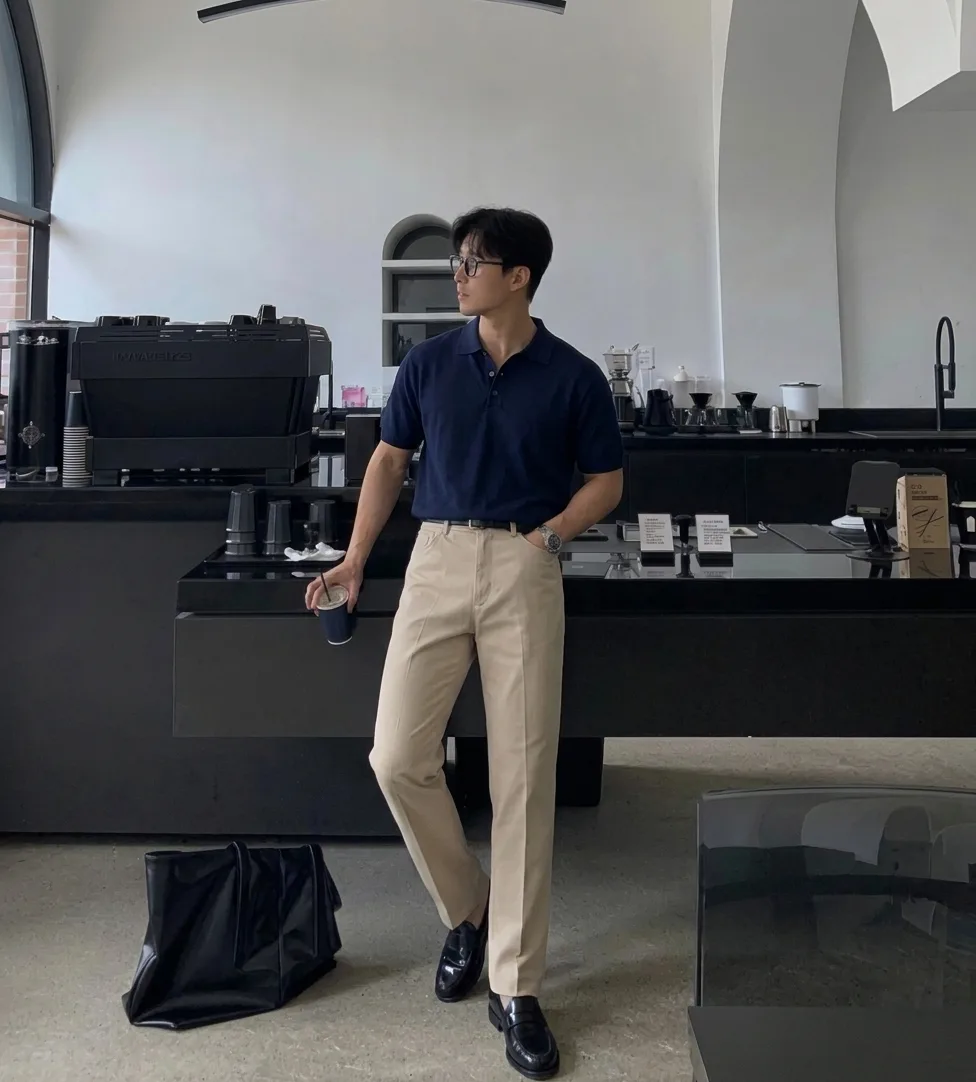 Man in a navy polo, beige trousers, and black loafers, holding a coffee in a cafe