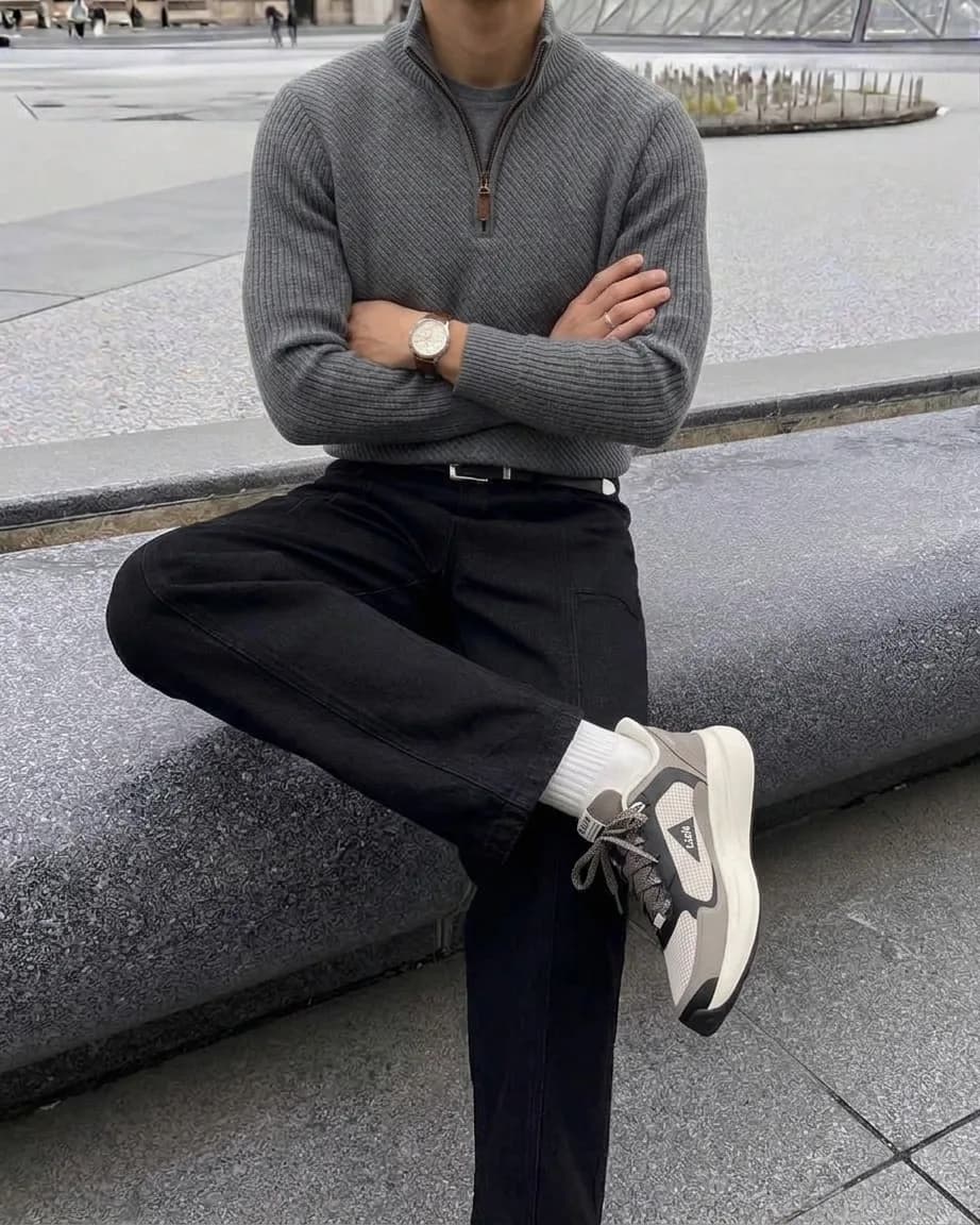 Man in a grey quarter-zip sweater, black jeans, and grey running shoes, seated on a stone bench