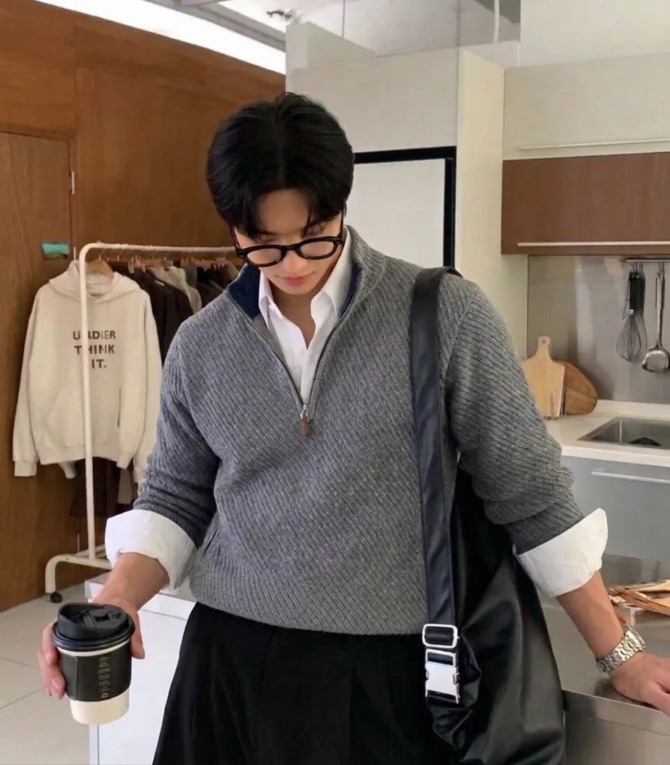 Man in a grey quarter-zip over a white button-down and black trousers, carrying a black bag and a coffee