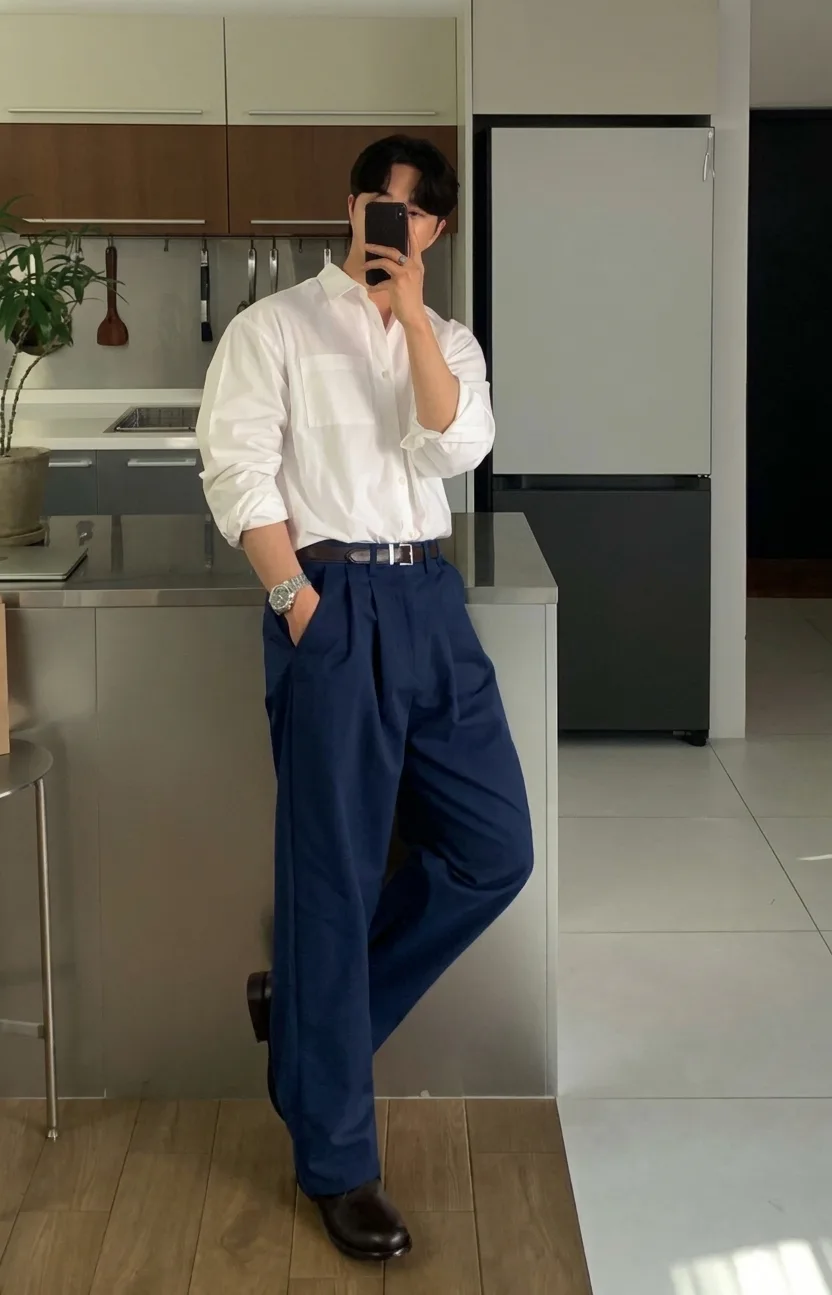 Man in a white button-down, navy pleated trousers, a brown belt, and brown shoes, taking a mirror photo in a kitchen