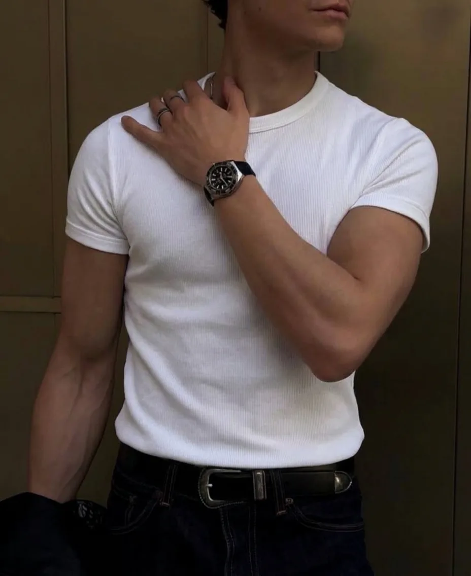 Man in a white ribbed crew-neck T-shirt and dark jeans, hand at his collar, with a metal watch and a black belt