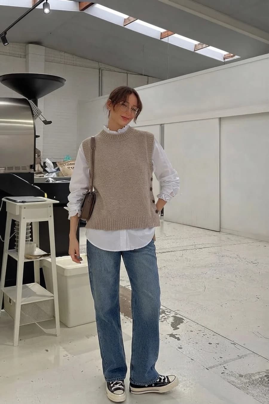 Women's winter outfit: cream knit vest over a ruffled shirt with wide jeans and sneakers